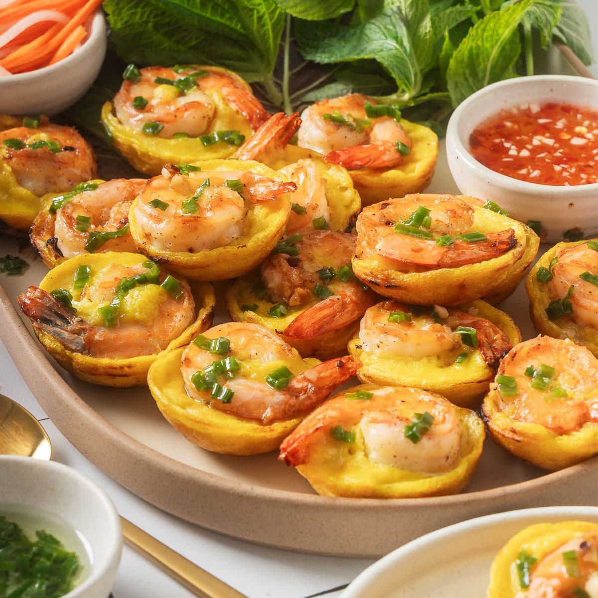 Đĩa bánh khọt vàng với nhân tôm, hành xanh, rau và chén nước chấm.